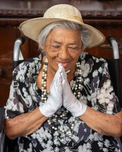 Elderly woman in a hat with hands in prayerful gesture, exuding grace.
