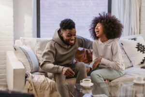 Cheerful young African American couple in casual outfit laughing happily while resting together on comfortable sofa at home