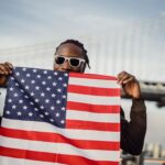 Young man with sunglasses holds the USA flag near Brooklyn Bridge, NYC.