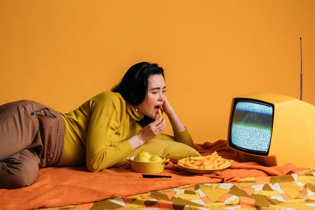 Woman dining on snacks while watching a vintage TV in a retro, colorful setting.