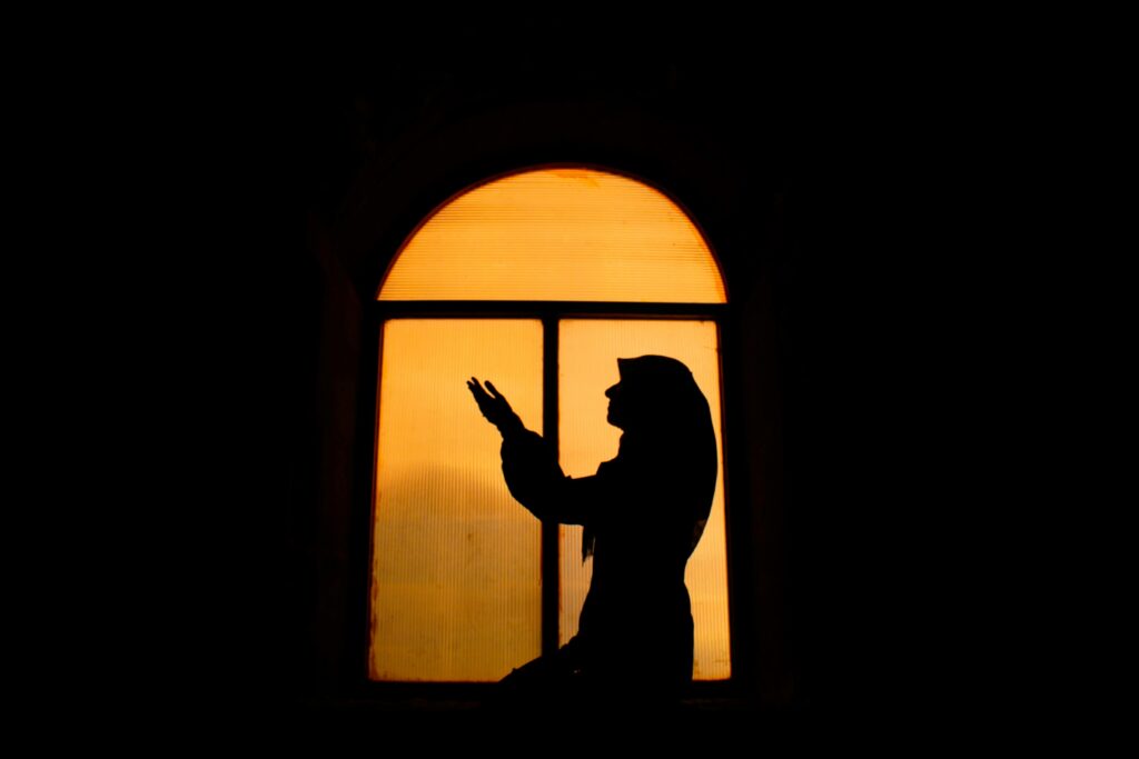 Silhouette of a woman in prayer by a window during twilight, creating a serene and reflective mood.