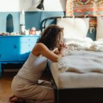 A woman kneeling by her bed in prayer, in a warmly lit, cozy bedroom setting.