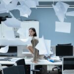 Woman perched on a desk in a chaotic office with papers flying, representing workplace stress.