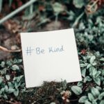 Close-up of an inspirational note with 'Be Kind' hashtag placed on lush green grass.