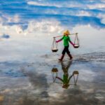 A worker carrying salt in baskets walks across a reflective water surface under a cloudy sky.