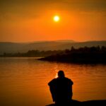 A person sits alone by a tranquil lake, with a stunning sunset in the background.