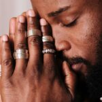 Close-up of a man praying with eyes closed, wearing rings on his fingers.