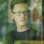View through wet window with crystal raindrops on thoughtful ethnic teenage man in eyeglasses looking away