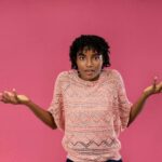 Young woman expressing confusion or uncertainty with a puzzled look on a pink background.