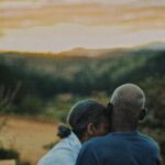 A loving elderly couple embraces while watching a scenic sunset in a natural setting.