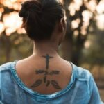 Back view of anonymous young female with Jesus Christ tattoo on back in casual clothes relaxing in park during sunset