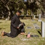 A man kneeling at a gravestone in a cemetery with an American flag.